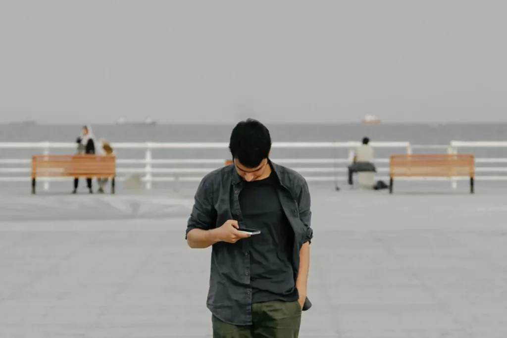 Image of a person looking down at their phone while standing with a grey background with two benches and people in the distance