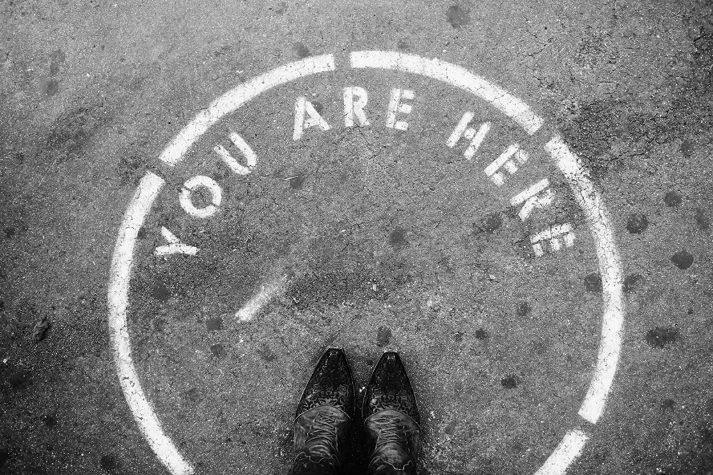 Feet with cowboy boots standing in the middle of a circle on pavement that says "you are here"
