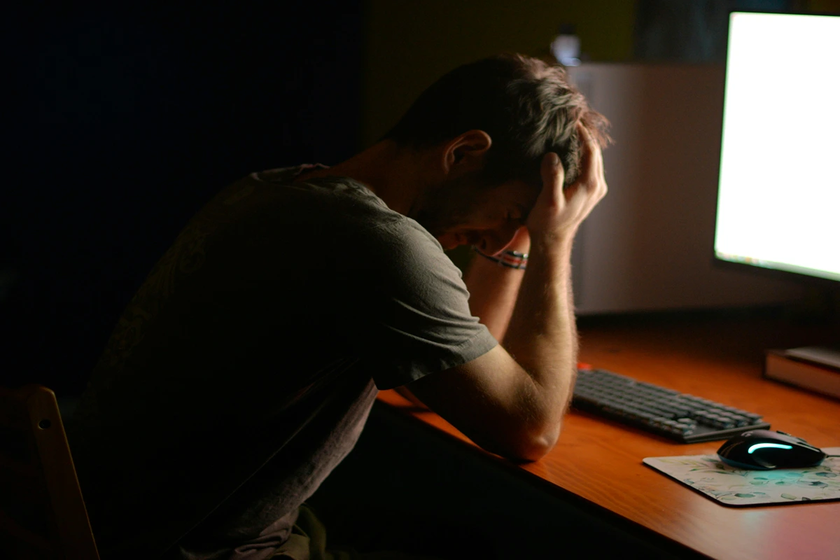 Image of person at their desk in a dark room with the computer light on and the person has their hands on their head