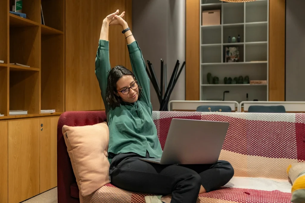 Image of person sitting on a couch inside with computer on their lap stretching their arms in the air