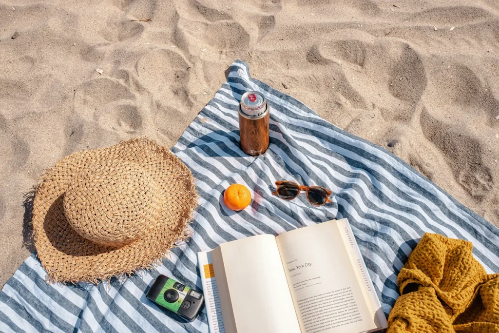 Image of white and great striped picnic blanket with a book, hat, drink, sunglasses, clementine and sweater on top of blanket