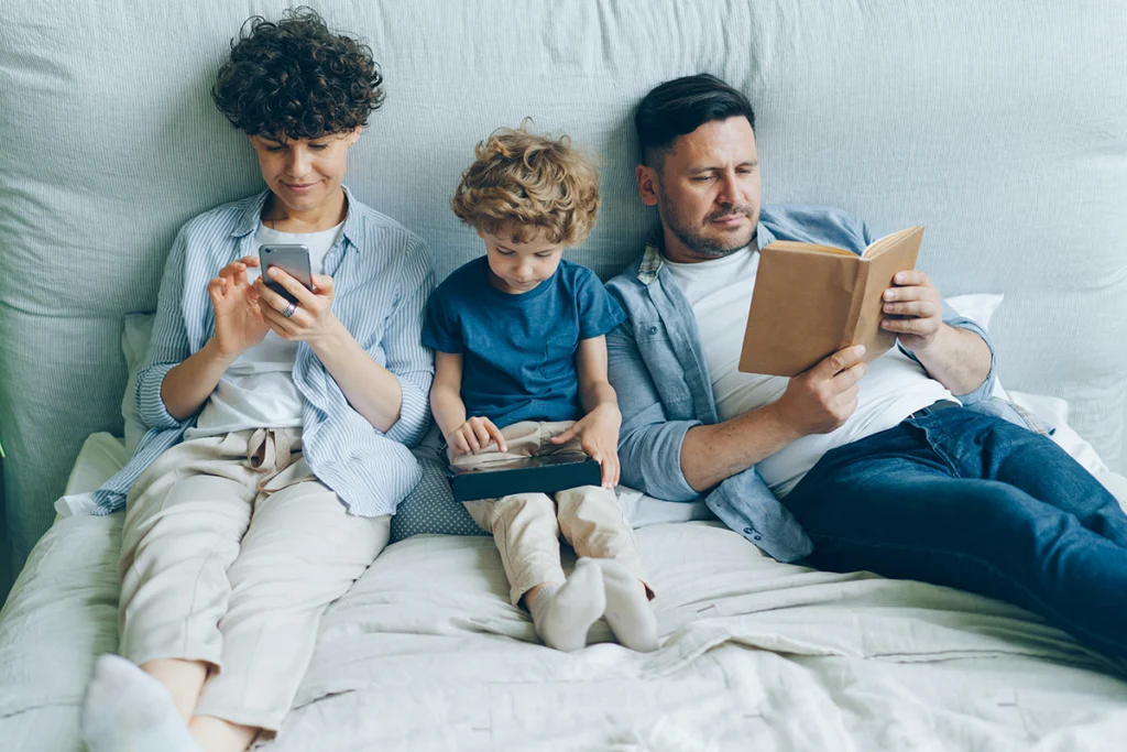 Image of a family in a bed with one looking at a cell phone, one at a book and the child looking at a screen