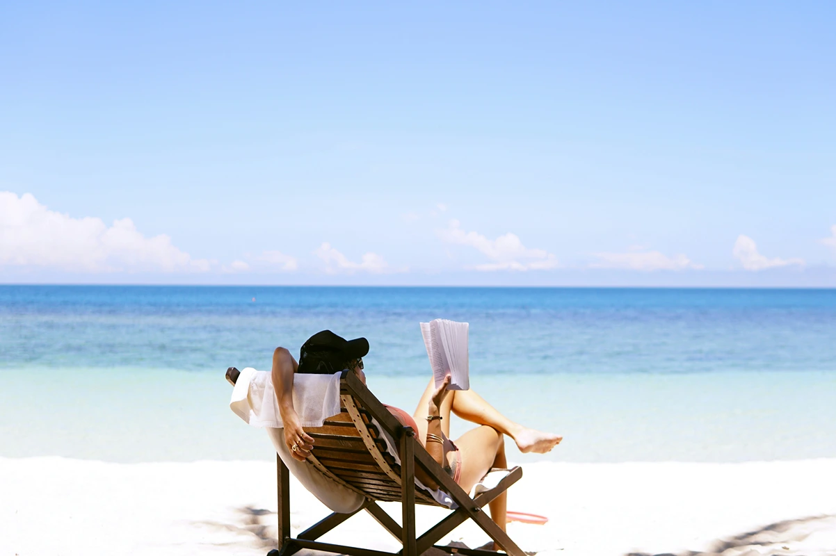 Image of a person on a sandy beach with blue water in the background reading a book relaxing