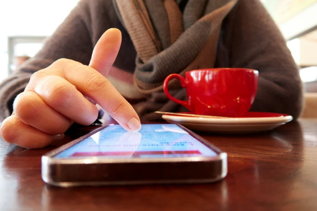 Close up photo of a cell phone and a person scrolling at a desk with a mug on it.