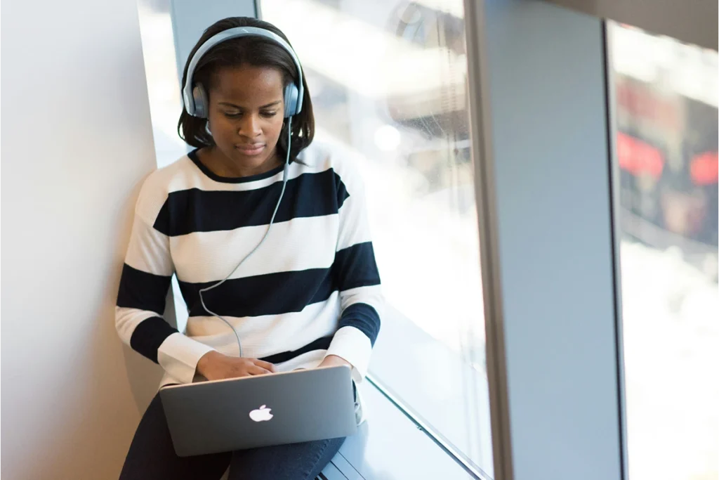 Image of person sitting at a window looking down at their computer with headphones on
