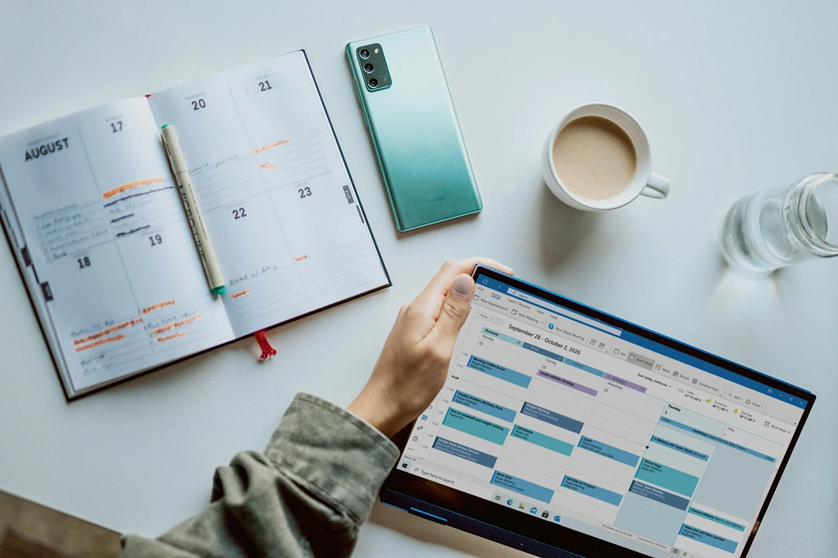 Desk with a persons hand looking at calendar on computer and written calendar.