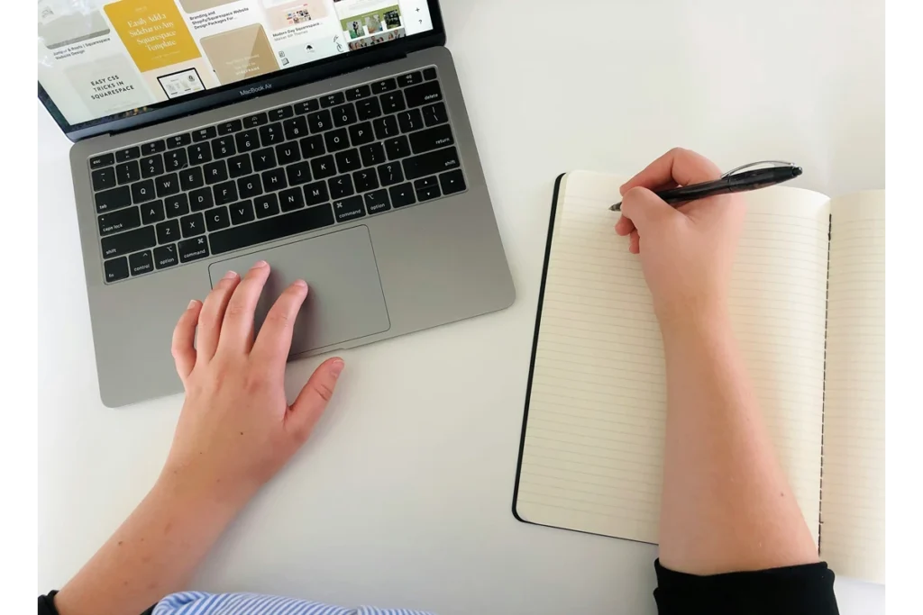 Image of hand writing in a notebook while multitasking on a computer