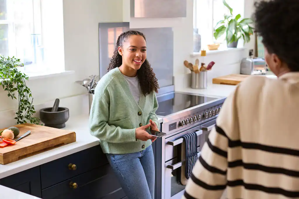 A parent and a teenager talking in a kitchen while the teenager holds a phone