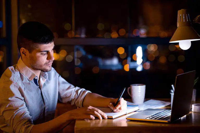 Man at a table at night under a lamp, writing in a notebook beside an open laptop.