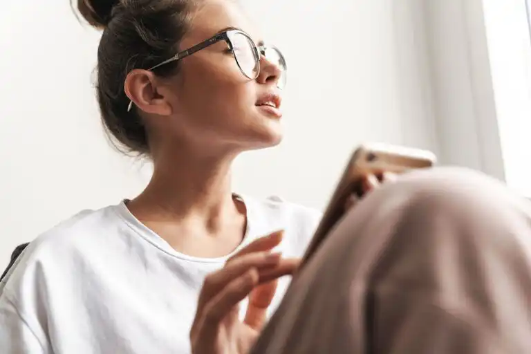 Woman sitting on a sofa in daylight holding her phone loosely in her lap and looking away from it.