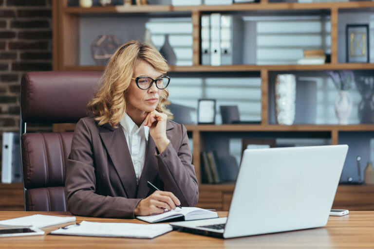 Woman working at a laptop at a desk with her phone face down at the far edge of the table.