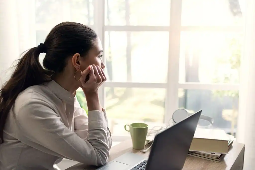 Woman sitting at a table by a window with a laptop in front of her, looking away from the screen.