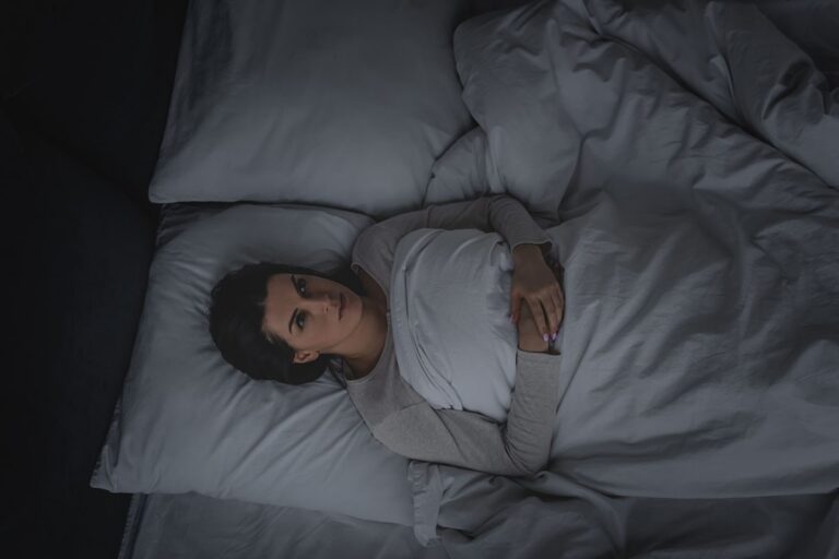 Woman lying awake in a dim bedroom at night, looking at the ceiling, with a hallway light through the door.