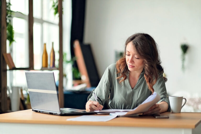 Woman working at a laptop with her phone face down on the desk beside her.