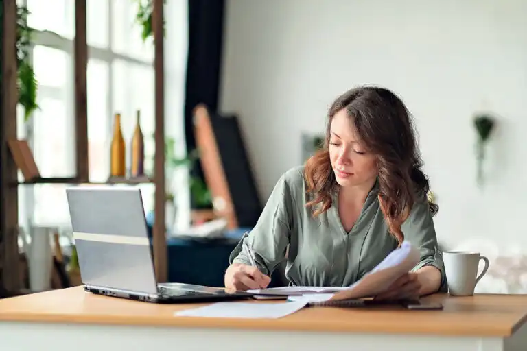 Woman working at a laptop with her phone face down on the desk beside her.