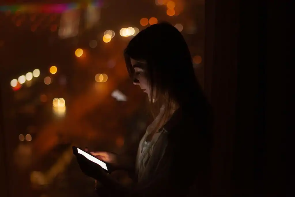 Woman standing at a window late at night, looking at her phone with the screen lighting her face.